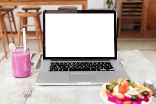 Modern portable laptop computer with blank white copy screen for your promotional text or advertisment, stands on table near delicious fruit salad and exotic fresh smoothie. Copy space on display