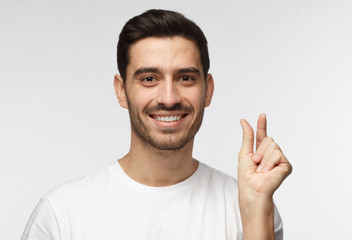 Fototapeta premium Young man with shows something tiny, smiling, shows white perfect teeth, isolated on grey background