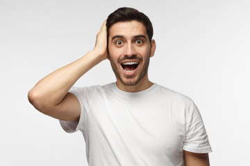 Headshot of handsome surprised man looking at the camera, astonished with big sale prices, holding hand on head, standing against grey background. Human face expression