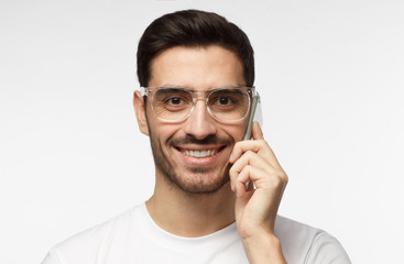 Closeup shot of young man in casual clothes isolated on grey background wearing stylish transparent eyeglasses enjoying telephone conversation with happy toothy smile laughing at joke from mate