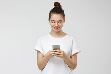 Naklejka premium Indoor shot of good-looking young woman isolated on gray background looking at smartphone, smiling openly while holding smartphone in both hands