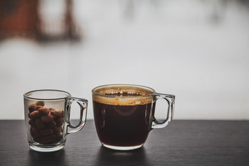 Transparent cup of coffee and a small glass cup filled with peanuts placed on dark brown laminated wooden surface in front of a window.