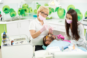 Obraz premium Attractive female dentist is smiling to the camera while holding medical instruments getting ready to attending the teeth of a male client of a dental office. Young brunette is assisting the doctor.