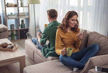Joyful married couple is using gadgets while drinking tea at home. They are sitting back to each other and smiling
