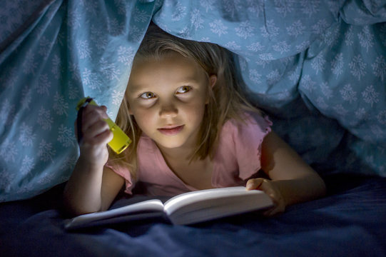 sweet beautiful and pretty little blond girl 6 to 8 years old under bed covers reading book in the dark at night with torch light smiling happy