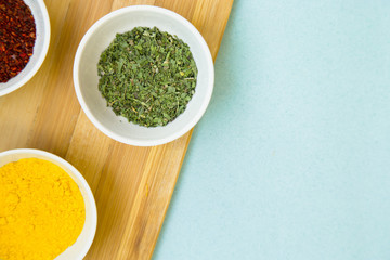 Plastic cups with dry spices and fresh herbs on a wooden cutting board with blue background, top view, close up
