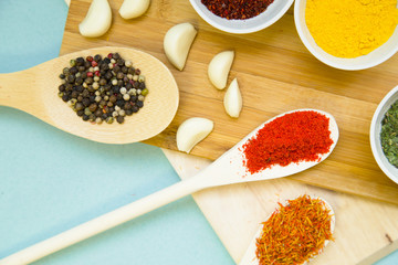 Plastic cups and spoons with dry spices and fresh herbs on a wooden cutting board with blue background, top view, close up
