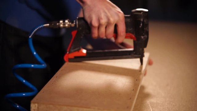 Man in a carpentry workshop using air pumped staple gun. He is stapling plywood along the straight line to the construction.