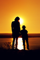 Silhouettes of mom and son holding hands and admiring the beautiful view of the sea on sunny evening at sunset time