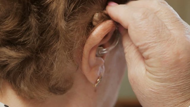 Close up of a senior woman inserting a hearing aid in her hear. Focus on the hearing aid