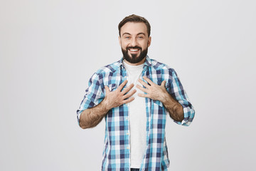 Portrait of pleased adult bearded male with sincere happy smile, holding hands on chest expressing surprise while standing over gray background. Employee is happy he was chosen to lead new project