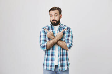 Portrait of puzzled bearded guy with lifted eyebrows showing confusion, crossing arms and showing in different directions with index fingers while standing over gray background.
