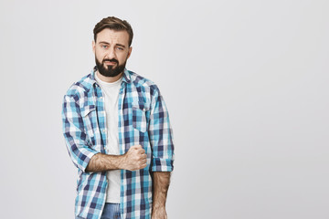 Fototapeta premium Attractive bearded european man with gloomy smile, squeezes fist, expressing tiredness and unwillingness to action, standing over gray background. Person is overloaded with work but should keep up