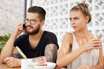 Irritated angry jealous young female being discontent as her bearded boyfriend speaks with unknown person via cell phone, doesn`t pay attention at her, hates disregard, sit together at coffee shop