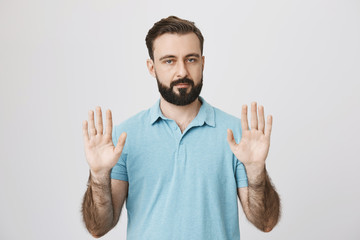 Portrait of calm adult man with beard raising his palms and looking at camera, standing over gray background. Guy suggests everyone to relax and breathe as team is under pressure.