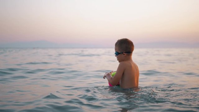 Slow motion shot of a child bathing in the sea in the evening. He floating on the ball which he taking to play