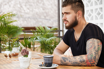 Pensive fashionable bearded male with tattoo, holds smart phone, enjoys online communication and wireless internet at outdoor cafeteria with tasty exotic beverage, thinks where to go on excursion