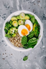 Healthy green vegetarian buddha bowl lunch with eggs, quinoa, spinach, avocado, grilled brussels sprouts and broccoli on dark grey background.