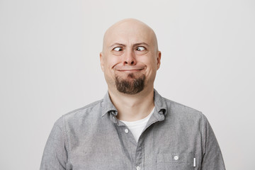 Obraz premium Portrait of funny and childish bald caucasian male model making crazy eyes and smiling strangely, standing over gray background. Guy shows daughter that he can see his own nose without mirror