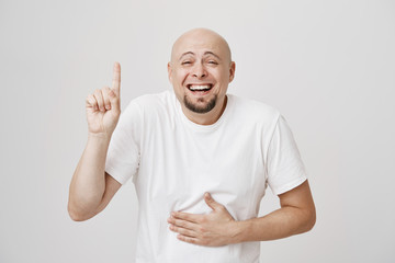 Close-up portrait of bald bearded guy pointing up and holding hand on belly while laughing out loud...