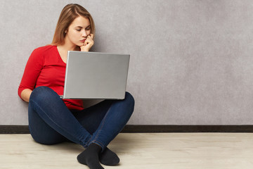 Carefree focused female dressed in red sweater and jeans, watches interesting movie on laptop computer with atttentive look, sits on floor, isolated over grey background. People, lifestyle concept