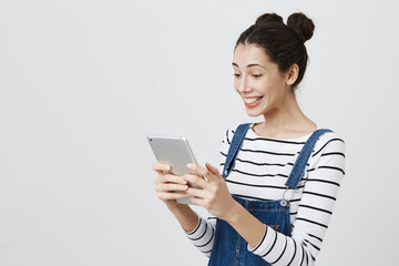 Fototapeta premium Half-turned european model with two buns hairstyle and pierced nose smiling cheerfully while holding tablet and browsing through it, standing in overalls against gray background.