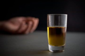 glass of whiskey or cognac or alcohol drink, hand of a drunk man in the background, alcoholism and alcohol abuse concept, defocused, selective focus, close up, gray table, dark background
