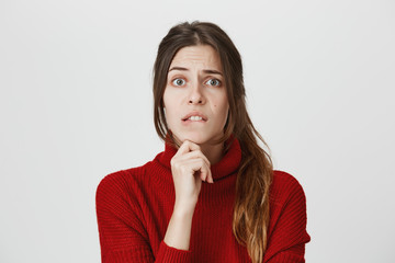 Portrait of young cute girl with brown hair who looks worried biting her lip and holding hand on chin, isolated over white background. Woman realised she made huge mistake in her report.