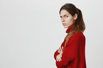 Profile of beautiful girl with ponytail in warm red sweater posing at studio, keeping arms folded,...