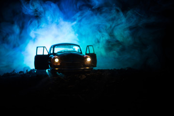 Silhouette of old vintage car in dark foggy toned background with glowing lights in low light, or...