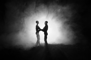 Love Valentine`s Day concept.Sillhouette of sweet young couple in love standing in the field and hugging on dark toned foggy background. Decoration with doll figures on table shot.