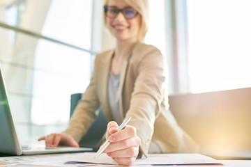 Cheerful young manager wearing eyeglasses looking at camera with toothy smile while distracted from work in spacious office with panoramic windows, focus on foreground