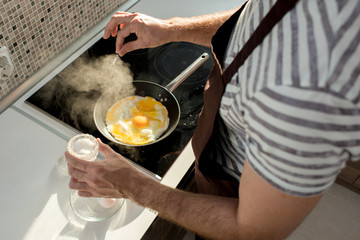 High angle close up of male hands salting eggs in frying pan while cooking breakfast in modern kitchen lit by sunlight