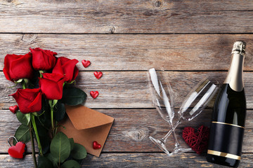 Champagne bottle with glasses, envelope and bouquet of red roses on wooden table
