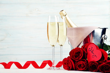Champagne bottle in bucket with glasses and bouquet of red roses on wooden table