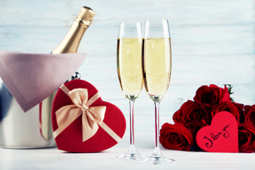 Champagne bottle in bucket with glasses, gift box and bouquet of red roses on wooden table