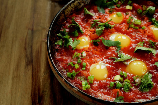 Fried eggs with tomato sauce and parsley in a cast iron pan on wooden background. Shakshuka a traditional meal of the Jewish cuisine