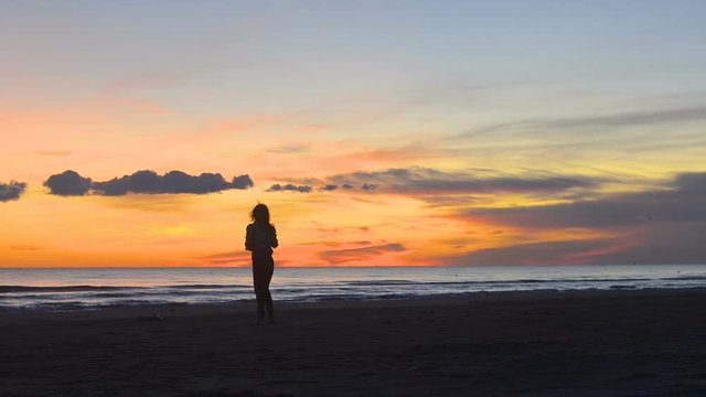  Pretty young woman walking happily along the beach at sunset. A video taken from a distance with a static image and the woman moves slowly from the camera and shows pleasure and happiness in the beau