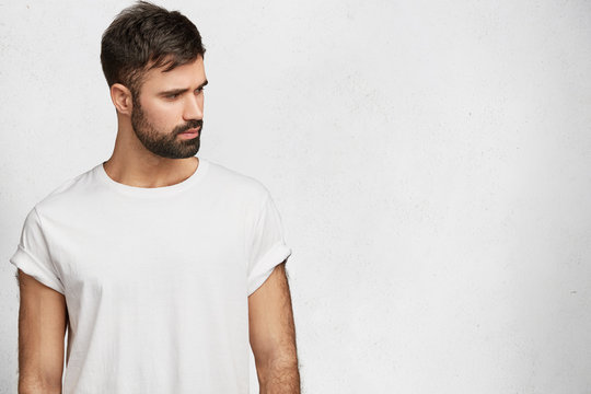 Clothing design and people concept. Serious bearded male dressed in casual white t shirt, looks pensively aside, isolated over white background with blank copy space for your advertising content