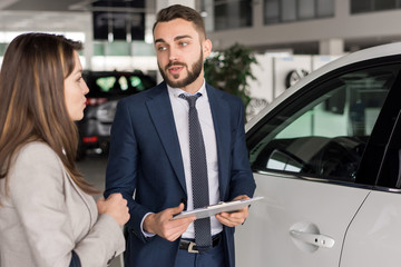 Fototapeta na wymiar Portrait of handsome car salesman talking to young woman in car showroom helping her choose