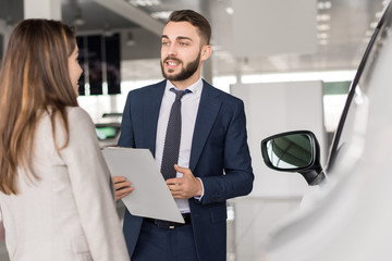 Fototapeta premium Portrait of handsome car salesman talking to young woman helping her choose in car showroom