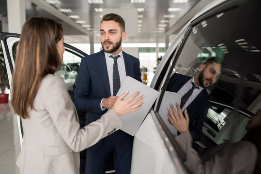 Portrait of handsome car salesman showing white luxury car to young woman in dealership showroom