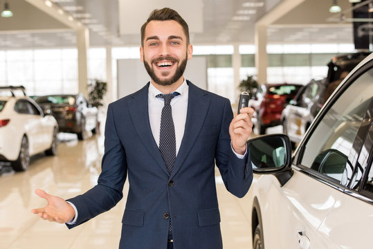 Portrait of handsome car salesman a holding car keys and smiling cheerfully at camera standing in luxury dealership showroom