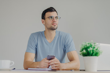 Thoughtful male freelancer works remotely on laptop computer, looks pensively and holds modern cell phone as waits for important call, isolated over grey background. Businessman poses indoor