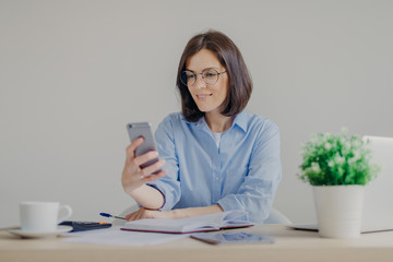 Successful brunette female freelancer recieves good news on smart phone while works on laptop computer, reads post on website, makes notes in diary, drinks strong coffee, makes some calculations