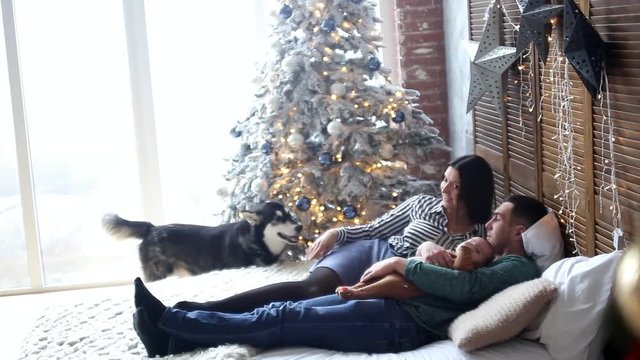 young couple with baby and dog lying on bed near Christmas tree