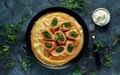 Crepes pancakes rolls with smoked salmon stuffed with wild rocket salad filling served on black plate with fresh creme.