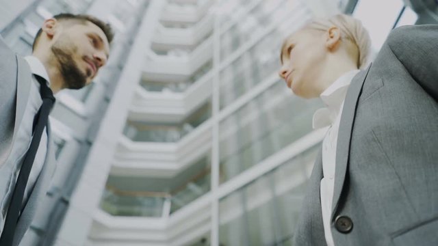 Low angle shot of bearded businessman shaking hands and talking with female business colleague in suit in hall of modern office building