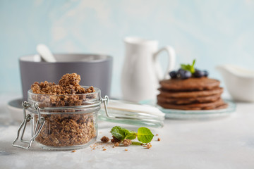 Chocolate baked granola in a glass jar, chocolate pancakes and milk. Healthy breakfast concept.