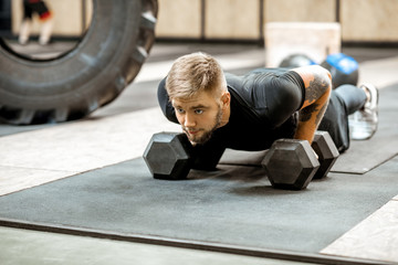 Athletic man in black sportswear with tattoos pushing ups on the dumbbells in the gym